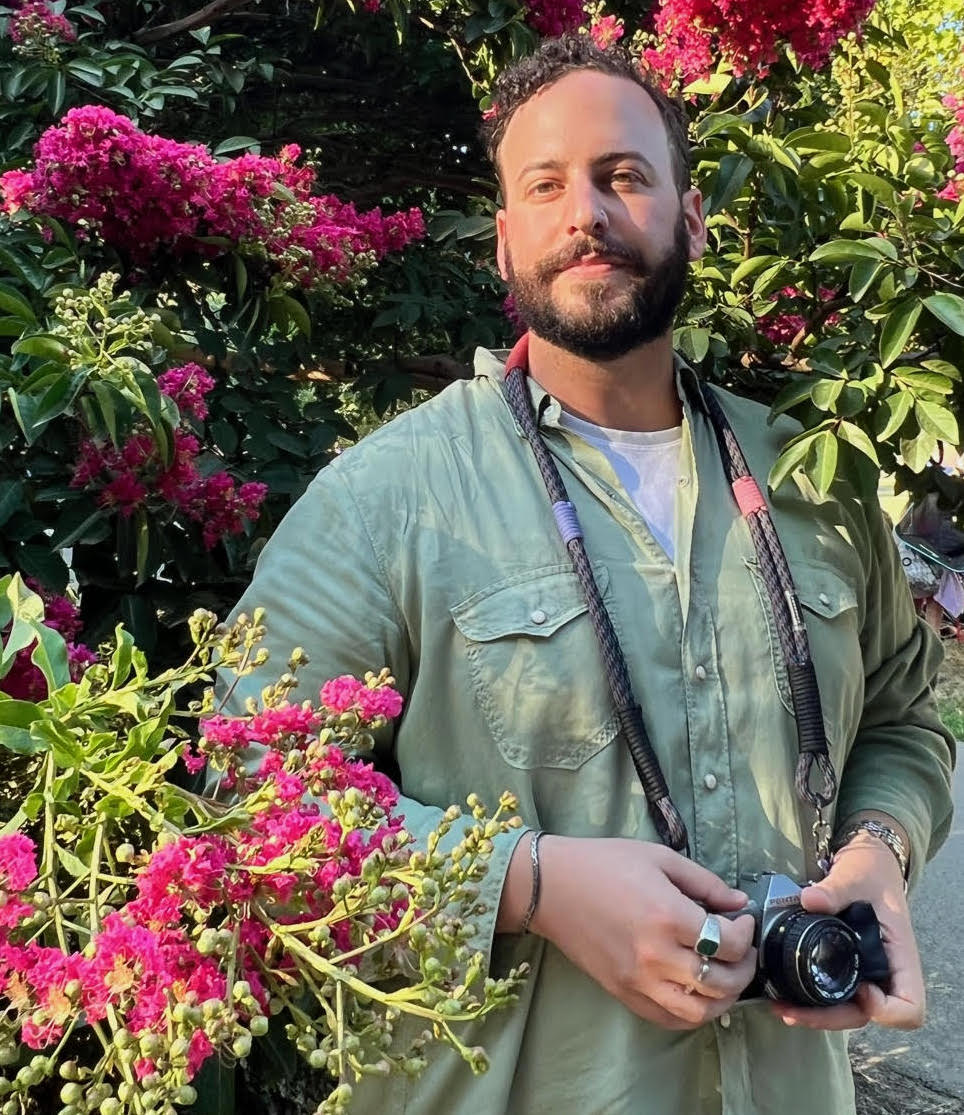 Mitch stands in some pink flowers with his camera around his neck